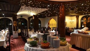 Un ristorante con tavoli rotondi apparecchiati per la cena, un'area buffet con cibo e bevande, e un'atmosfera calda e soffusa con luci a stringa.