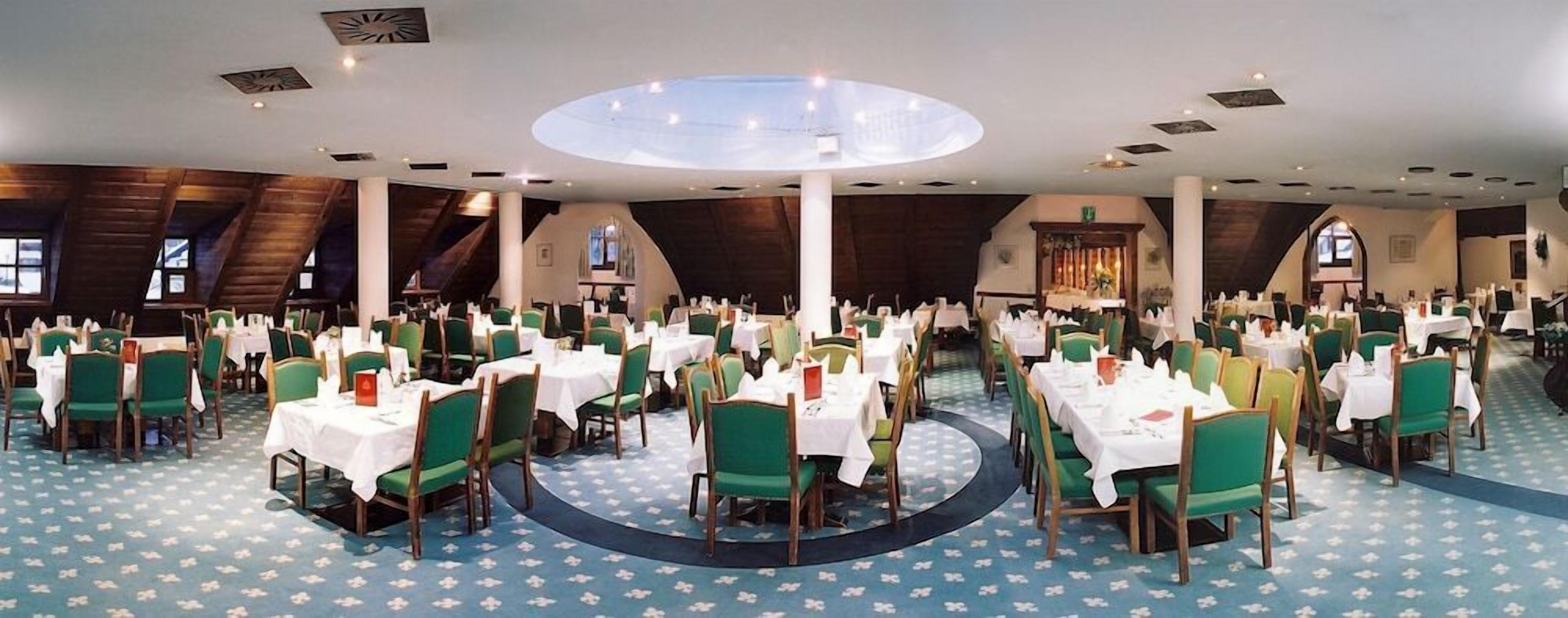A large dining hall with multiple round tables and green chairs, arranged in a spacious layout with high ceilings and a central skylight.