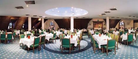A large dining hall with multiple round tables and green chairs, arranged in a spacious layout with high ceilings and a central skylight.