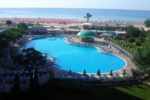 A large, curved swimming pool surrounded by lounge chairs and umbrellas, with a beach and ocean in the background. - Hotel Slavyanski (Nessebar)