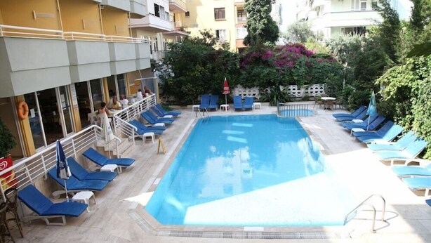 A swimming pool with blue lounge chairs and a small table, surrounded by greenery and a building with balconies. - Alin Hotel (Alanya)