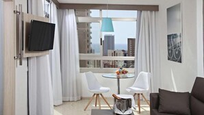 A modern hotel room with a large window, a flat-screen TV mounted on the wall, a small round table with two white chairs, and a brown sofa. - Flamingo Beach Resort (Benidorm)