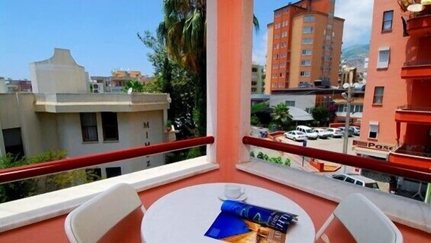 A balcony with a table and chairs, overlooking a street with parked cars and buildings. - Caprice Apartments (Alanya)