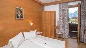 A bedroom with a wooden headboard, a bed with white linens, a wooden wardrobe, a chair, and a view of mountains through a window with floral curtains. - Guesthouse Schneeberger (Zell am Ziller)