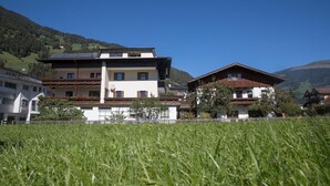 A multi-story building with a flat roof and a balcony, surrounded by a grassy field and a mountain backdrop. - Guesthouse Schneeberger (Zell am Ziller)