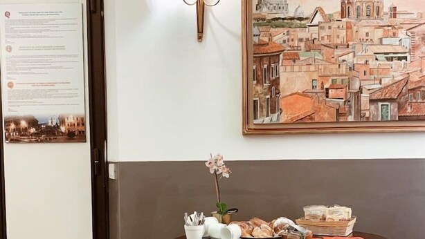 A room with a dining table set with plates, a vase with flowers, and a lamp. There is a painting of a cityscape on the wall. - Residenza Sciarra (Rome)