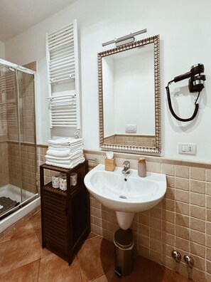 A bathroom with a white sink, a mirror with a decorative frame, a hairdryer on the wall, and a stack of towels on a wooden shelf.
