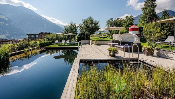 A serene outdoor pool area with lounge chairs and umbrellas, surrounded by lush greenery and a mountain backdrop. - Crystal Hotel (Fuegen)