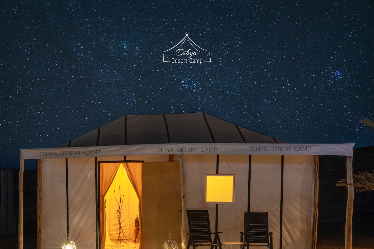A desert camp with a tent labeled "Dihya Desert Camp," a pathway leading to the entrance, and two chairs outside.