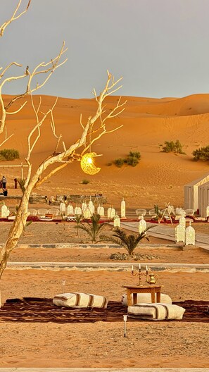 A desert setting with a dining area featuring white chairs and tables, a tree with bare branches, and a decorative hanging light. - Dihya Desert Camp (Taouz)