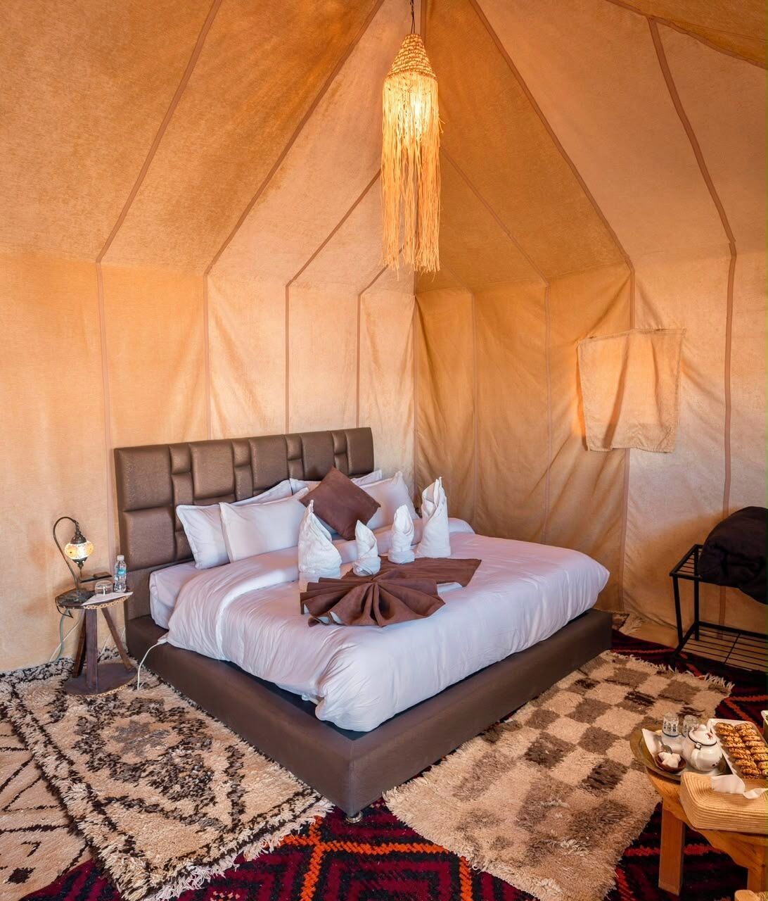 A tent-style room with a bed, a hanging light fixture, a bedside table with a lamp, and a small table with a tray of items.