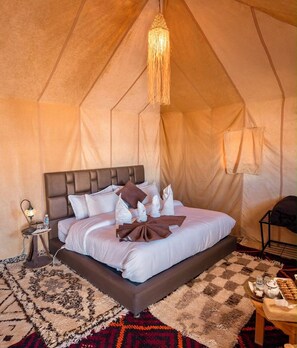 A tent-style room with a bed, a hanging light fixture, a bedside table with a lamp, and a small table with a tray of items. - Dihya Desert Camp (Taouz)
