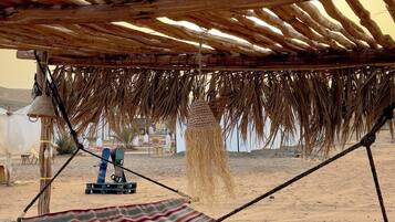 A beach hammock under a wooden pergola with a striped blanket, a water pump, and a thatched roof.