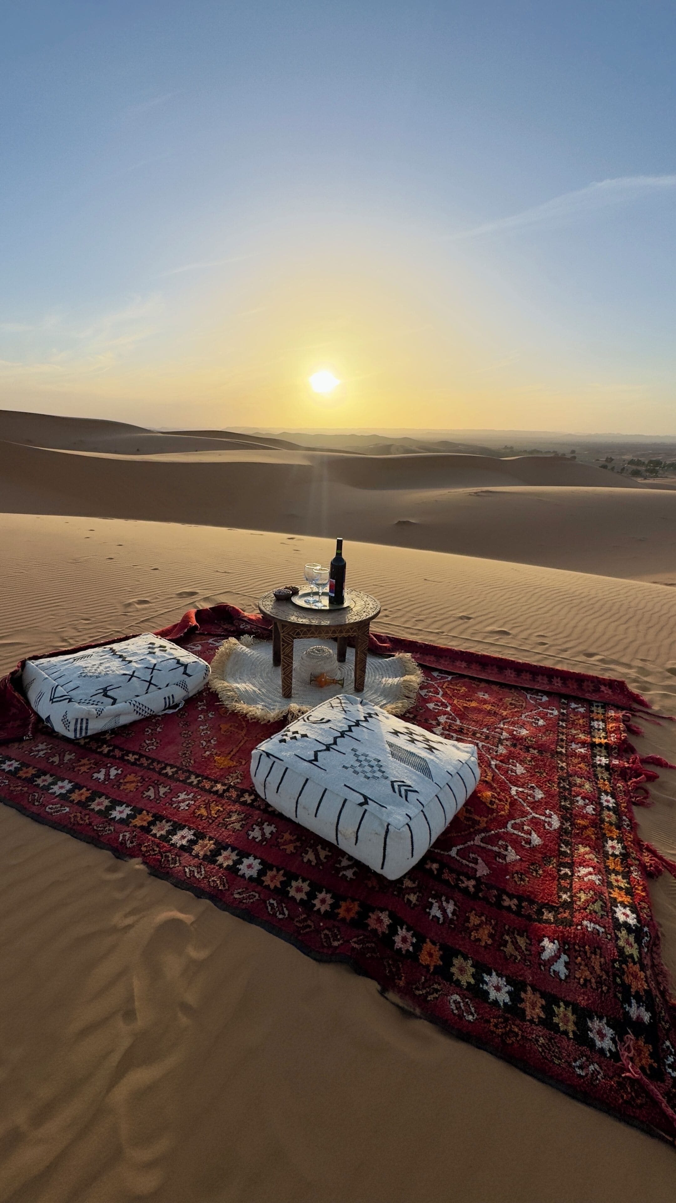 Escena desértica con una manta y almohadas sobre la arena, una pequeña mesa con una botella y vasos, y el sol poniéndose al fondo.