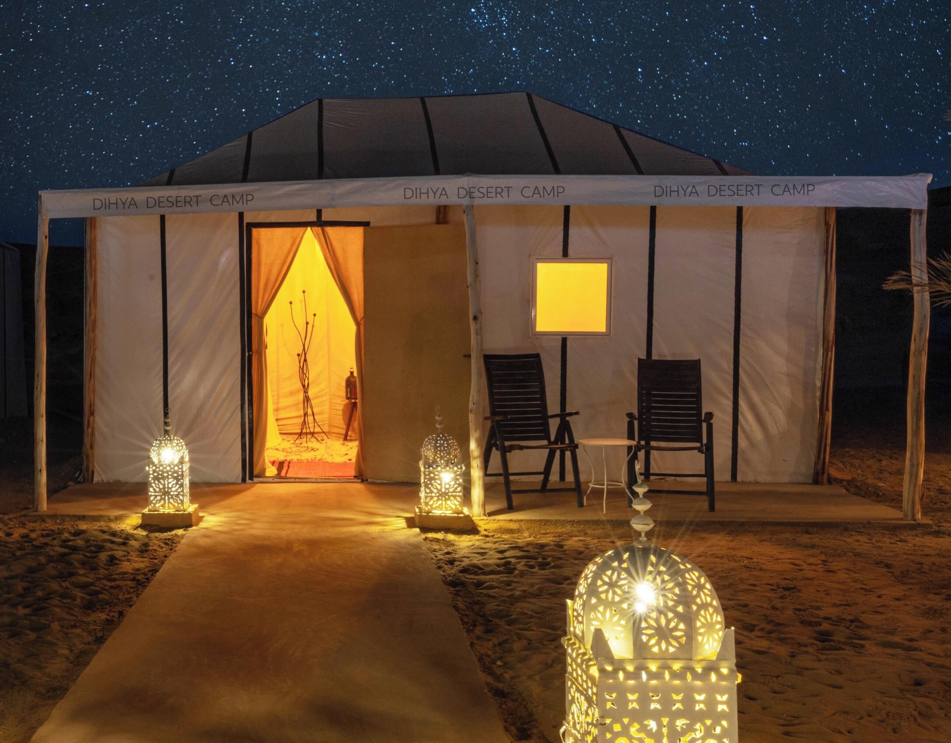 Un campamento en el desierto con la entrada de una tienda, dos sillas y faroles decorativos.