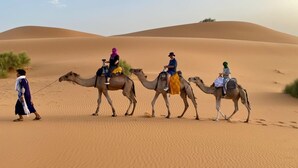 Un grupo de personas montando camellos a través de un paisaje desértico con dunas de arena al fondo.