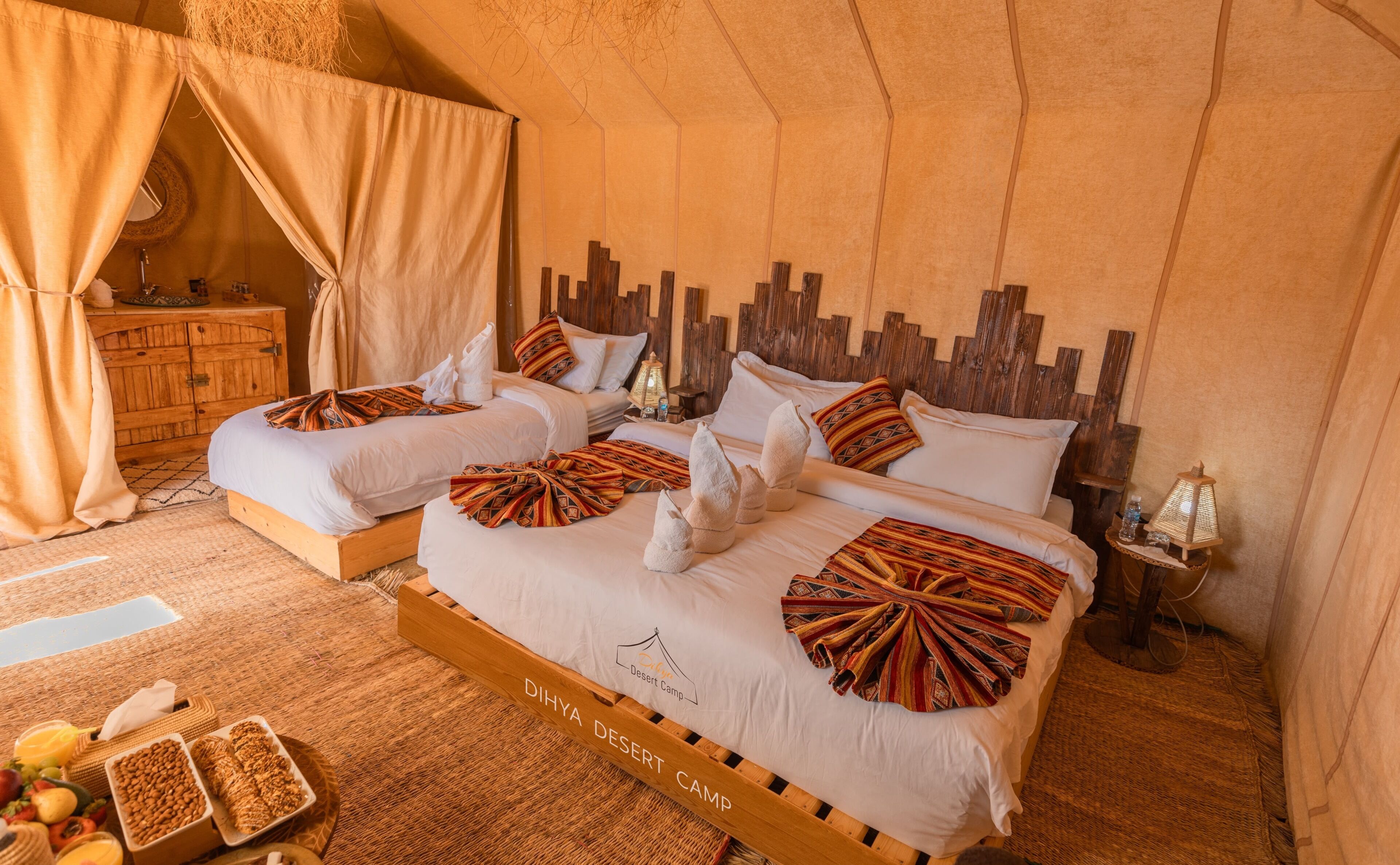 A room with two beds, a wooden headboard, a woven rug, and a small table with a lamp.