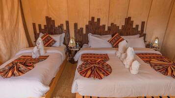 A hotel room with two beds, each with a towel and a patterned blanket. There are two bedside tables with lamps and a bottle of water.