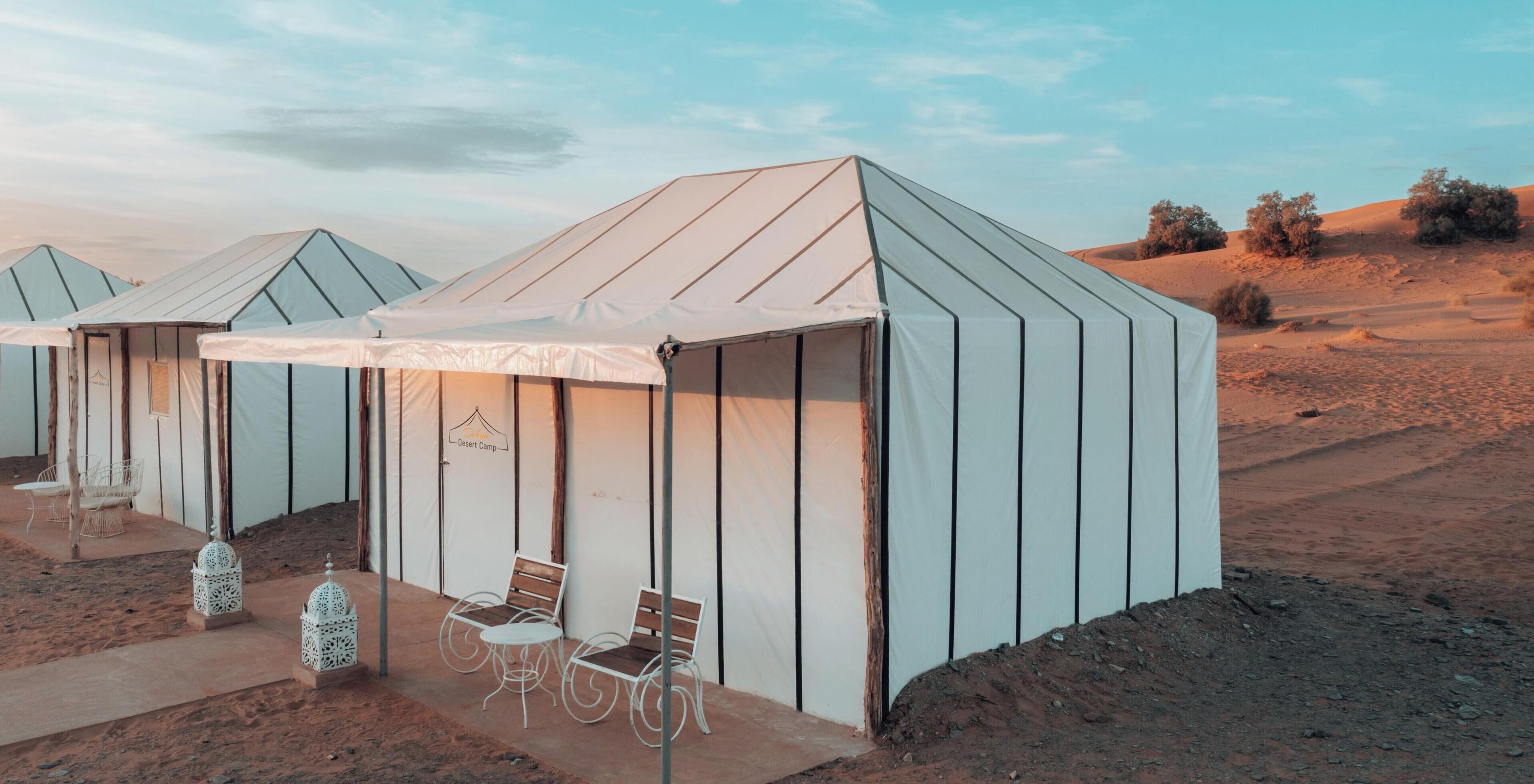 Acampamento no deserto com tendas, cada uma com um banco e uma mesa.