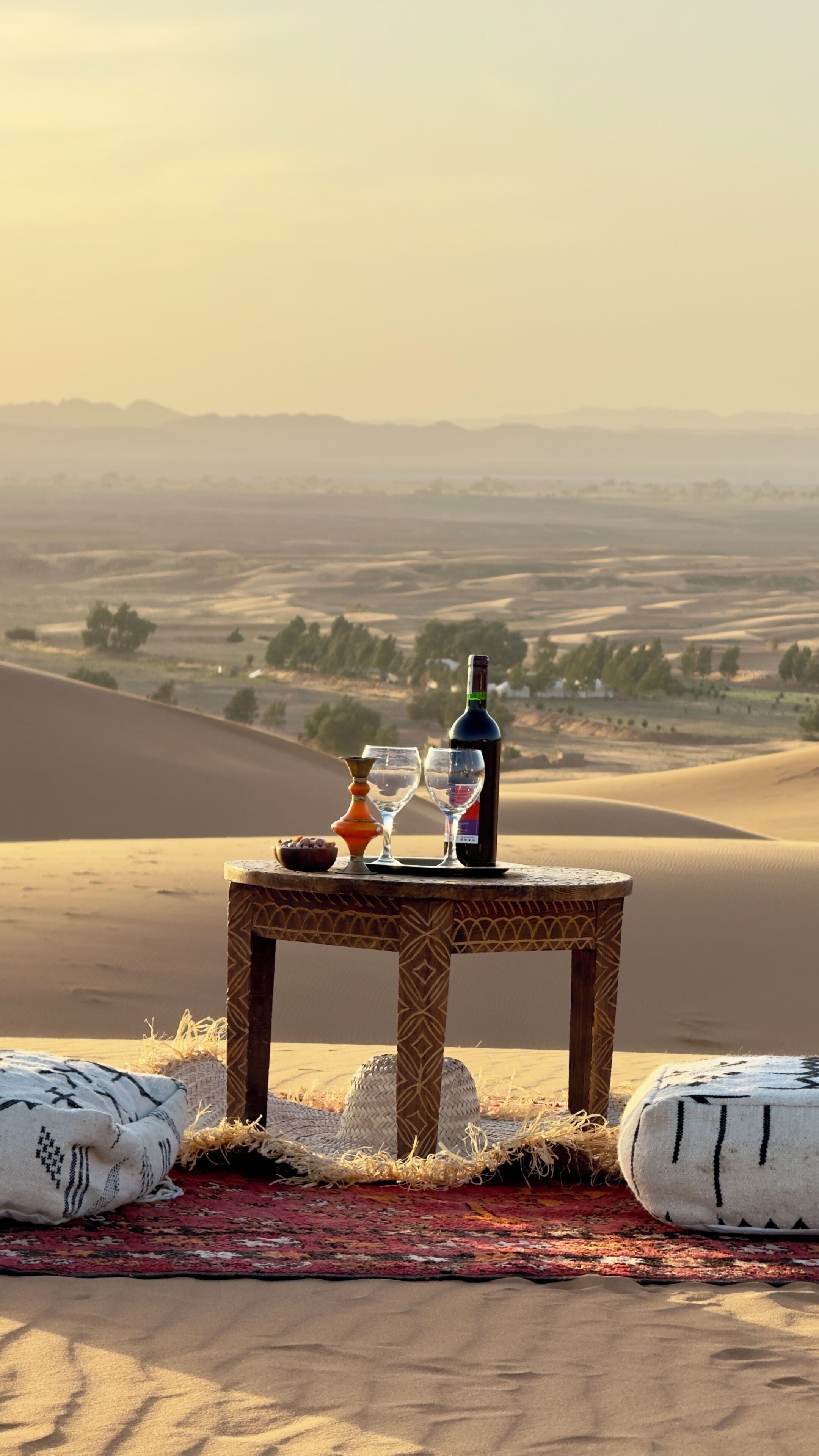 A table with a bottle and glasses on a woven mat, set against a backdrop of sand dunes and distant hills.