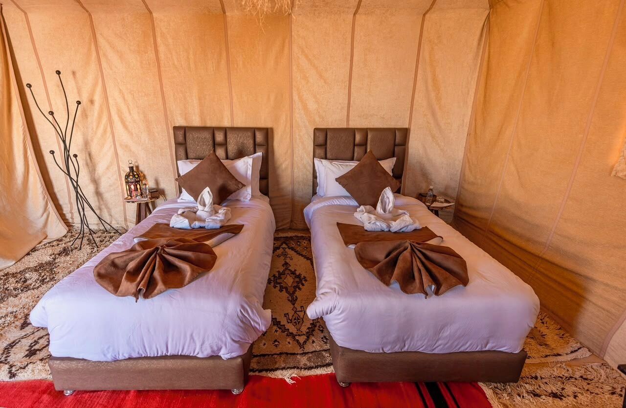 Habitación con dos camas, cada una con cabecera, almohadas y manta. Hay una mesita con lámpara y un objeto decorativo.