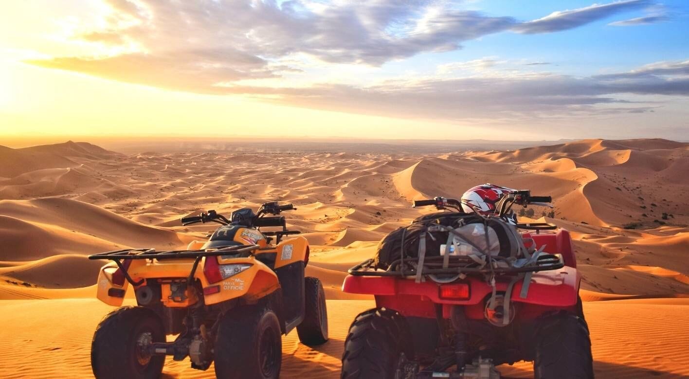Two ATVs parked on a desert landscape with sand dunes in the background.