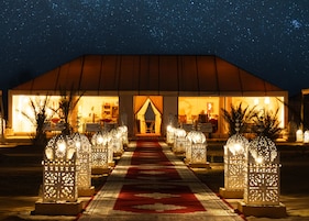 Un campamento en el desierto con un cielo estrellado, una alfombra roja que lleva a una estructura tipo tienda, y faroles ornamentales a lo largo del camino.