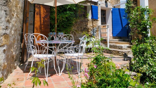 Une terrasse extérieure avec des chaises et une table en métal blanc, une pergola avec des volets bleus et un mur en pierre.