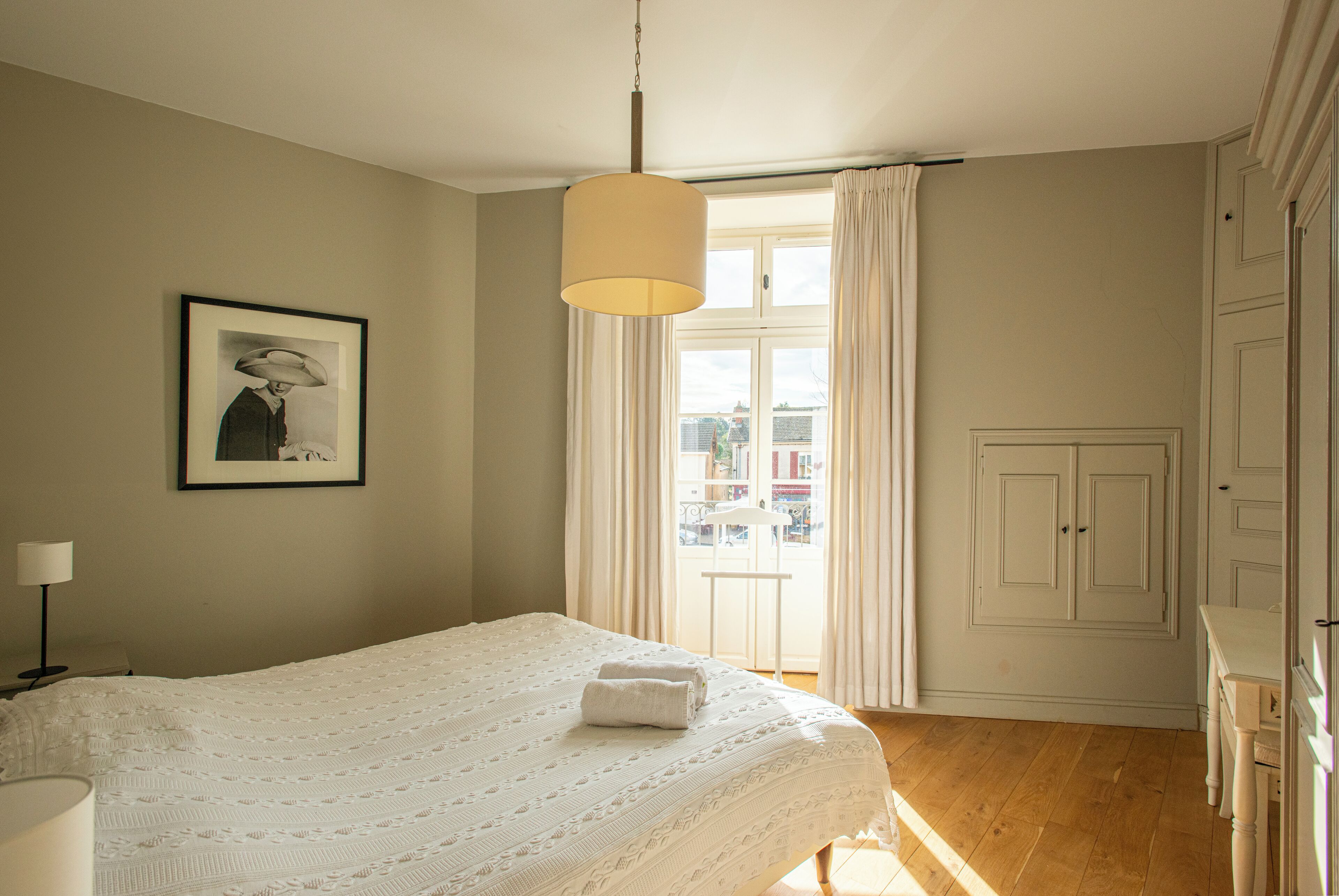 A bedroom with a bed, a hanging light fixture, a framed picture on the wall, a window with curtains, and a door.