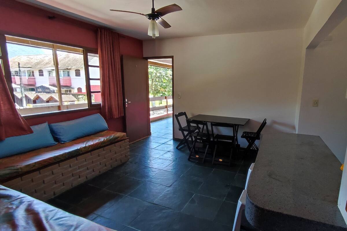 Una habitación con sofá, una mesa y sillas. Se ve una ventana con cortinas, una puerta y un ventilador de techo.