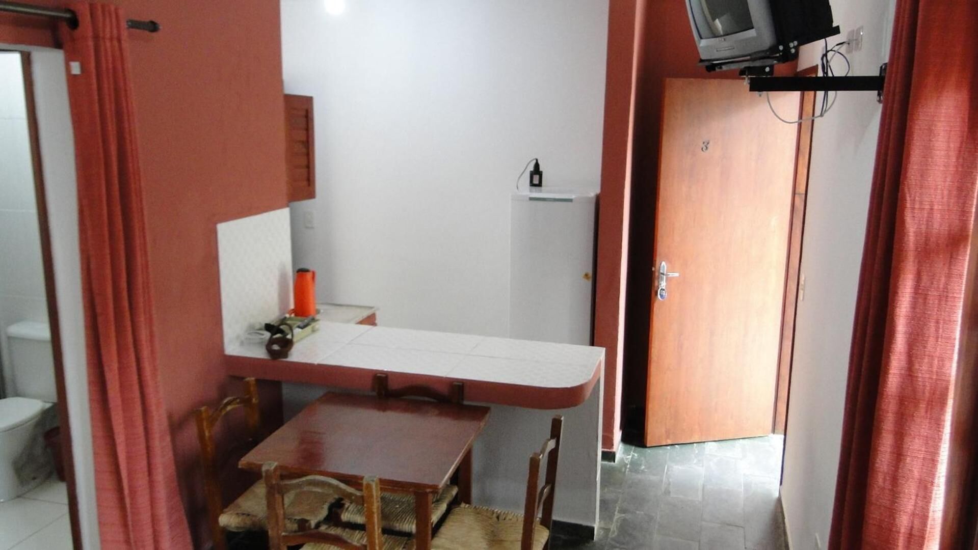 Een kleine kamer met een houten tafel, een stoel, een aan de muur bevestigde televisie en een deur met het nummer '3'.