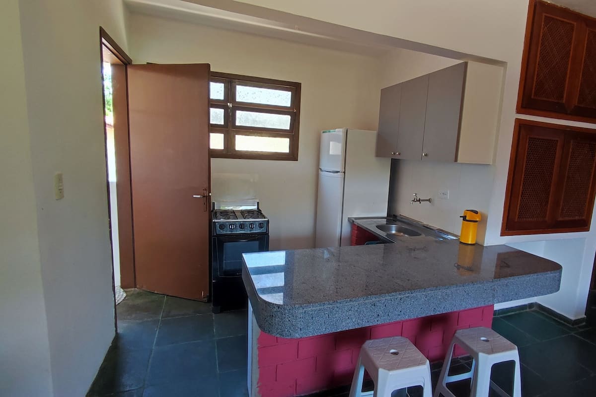 Cocina compacta con estufa a gas, refrigerador y pileta. Hay dos taburetes y un basurero pequeño.