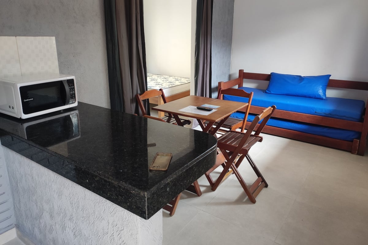 Una habitación compacta con kitchenette, una mesa de comedor con sillas, un sofá azul y una ventana con cortinas.