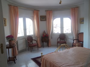 Een slaapkamer met een bed, twee stoelen, een kleine tafel en een raam met gordijnen.