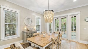 A dining room with a wooden table, wicker chairs, a chandelier, and a view through large windows. - Pet-Friendly WaterColor Retreat | Pool Spring '27 | Cart & Bikes (Santa Rosa Beach, Florida)