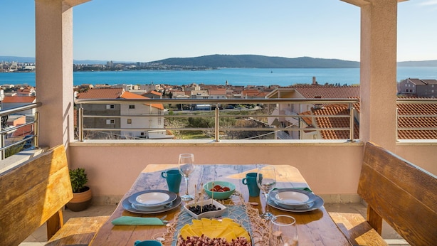 A balcony with a table set for two, overlooking a coastal cityscape with a view of the sea and hills.