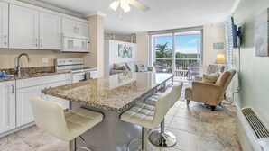 A kitchen with white cabinets, a granite countertop, and a ceiling fan. There is a bar area with bar stools and a view of the outdoors. - Beach & Tennis 1301 (Bonita Springs)
