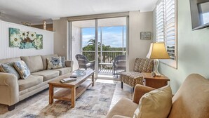 A living room with a sofa, coffee table, and chairs, a balcony with a view, and a welcome sign. - Beach & Tennis 1301 (Bonita Springs)