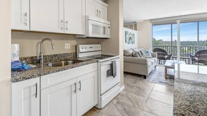 A modern kitchen with white cabinets, a stainless steel sink, and a microwave. There is a dining area with a table and chairs, and a balcony with a view. - Beach & Tennis 1301 (Bonita Springs)