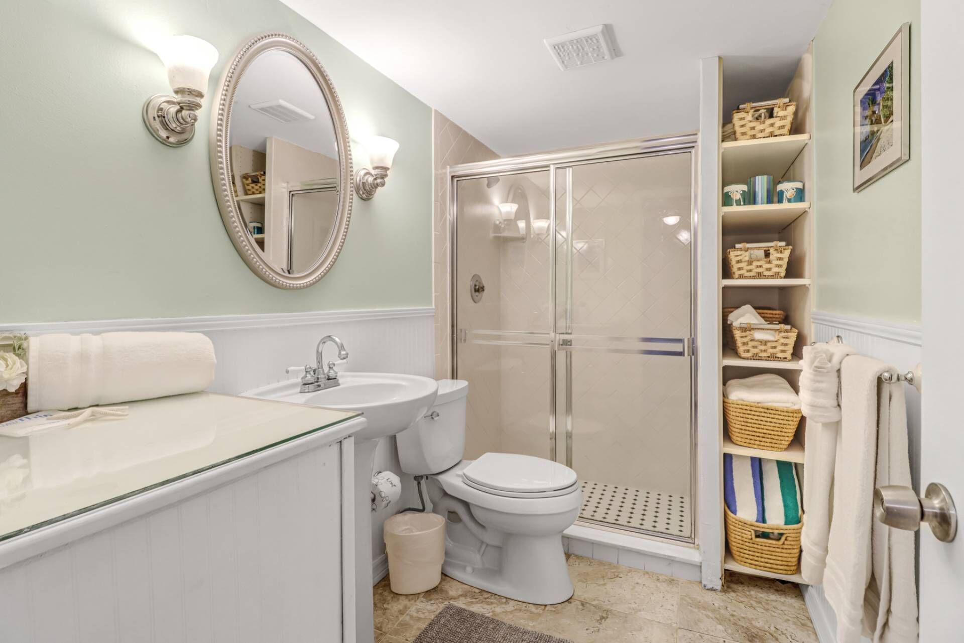A bathroom with a glass shower enclosure, a white sink, and a toilet. There are wicker baskets on shelves and a mirror with a decorative frame.