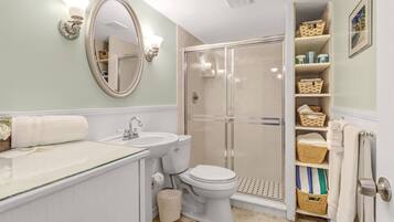 A bathroom with a glass shower enclosure, a white sink, and a toilet. There are wicker baskets on shelves and a mirror with a decorative frame.