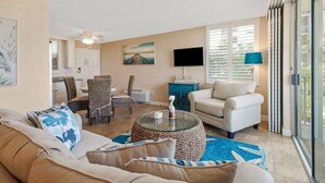A living room with a sofa, armchairs, a coffee table, and a TV. There is a painting on the wall and a window with blinds. - Beach & Tennis 3204 (Bonita Springs)