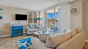 A living room with a beige sofa, a blue patterned rug, a glass coffee table, and a television on a stand. - Beach & Tennis 3204 (Bonita Springs)