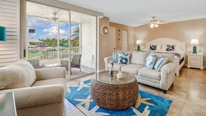 A spacious living room with a balcony, a round woven coffee table, a white sofa with blue and white cushions, and a white headboard bed. - Beach & Tennis 3204 (Bonita Springs)