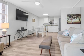 Un salon moderne avec un canapé, un banc et un téléviseur. On aperçoit au fond une cuisine avec des meubles blancs et un coin repas.
