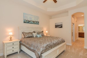 A bedroom with a large bed, two bedside tables with lamps, a ceiling fan, and artwork on the walls.