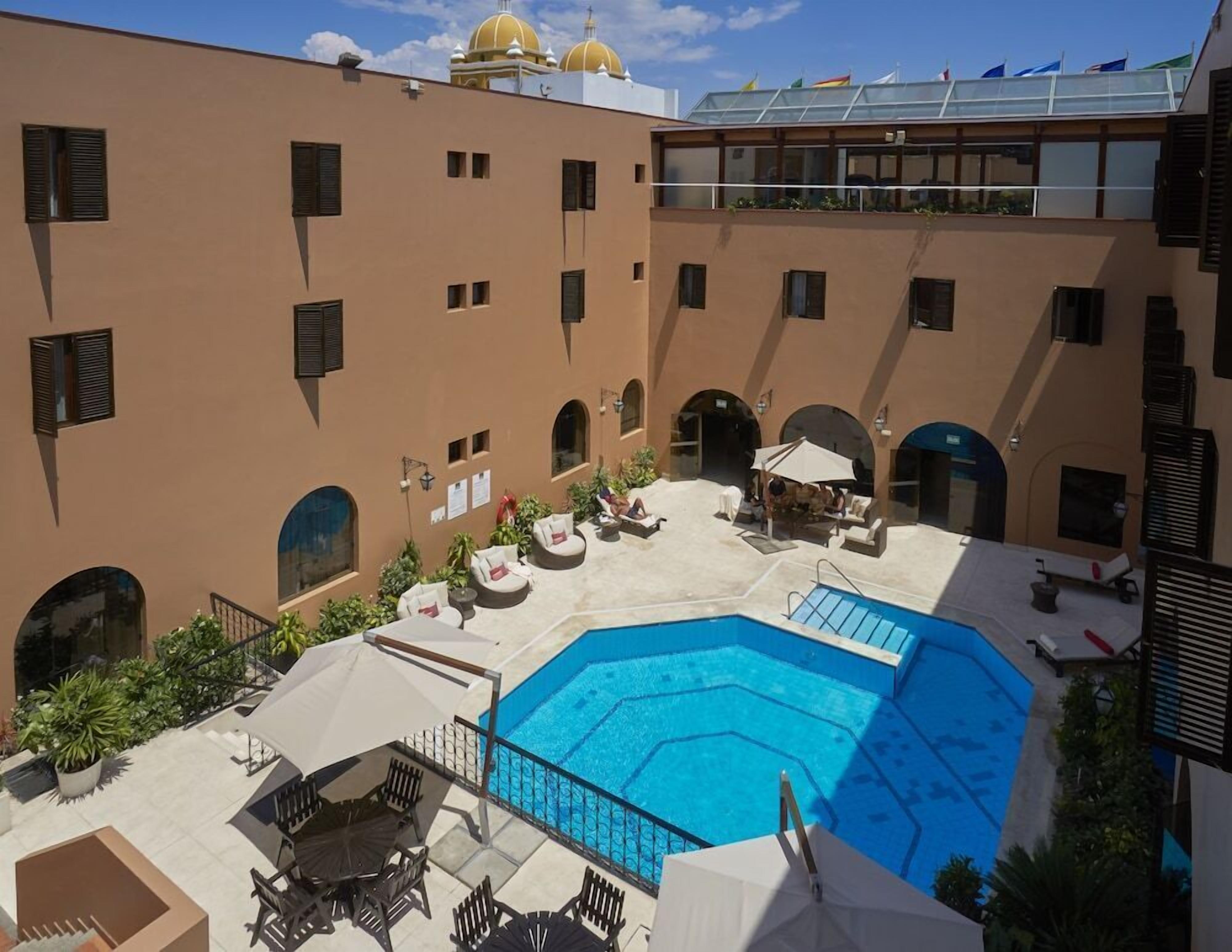 A rooftop pool area with lounge chairs and umbrellas, surrounded by a building with multiple balconies and arched doorways.