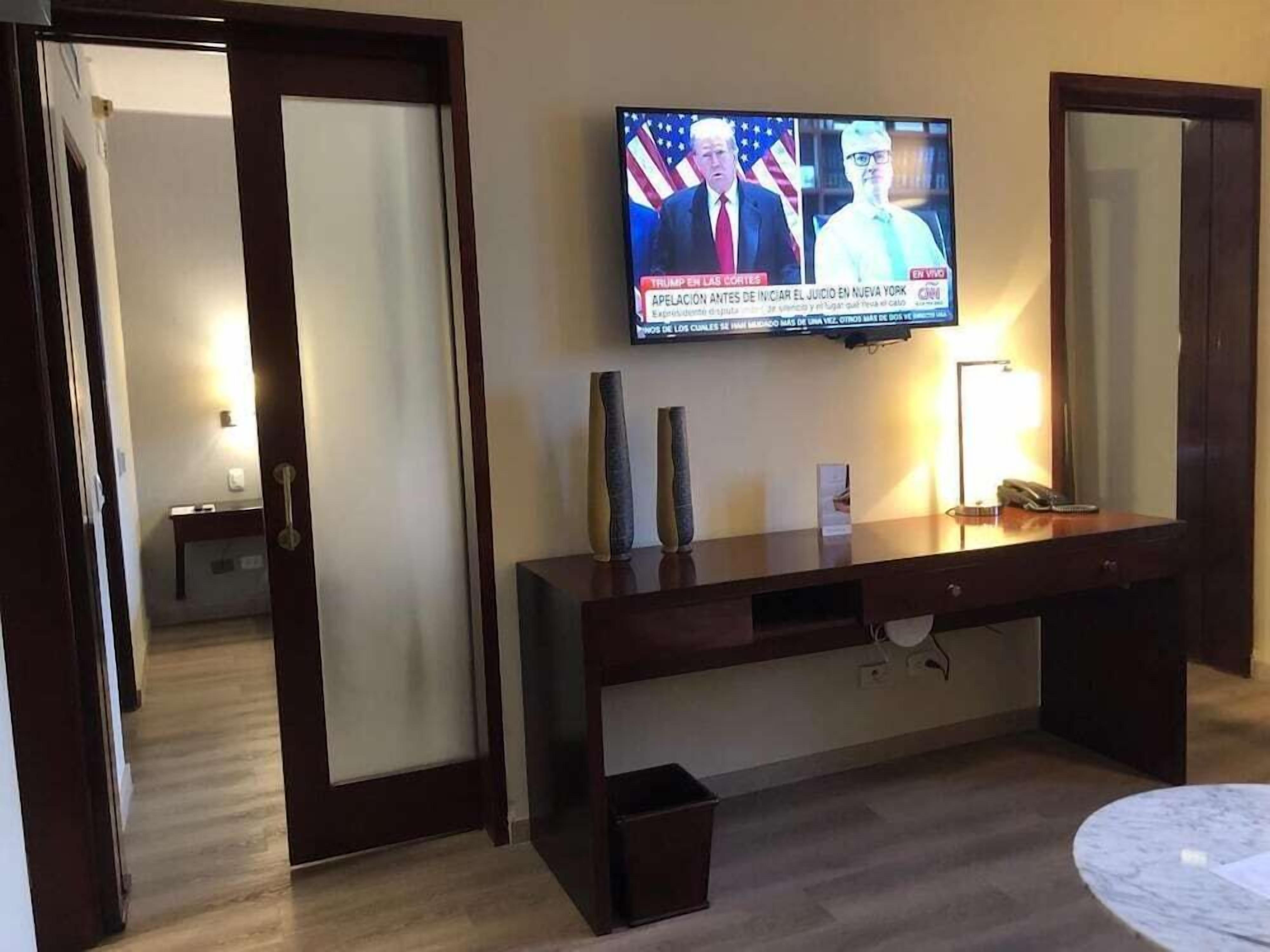 Habitación de hotel con una televisión montada en la pared, un escritorio de madera, una mesita de noche con lámpara y un pequeño cesto de basura.