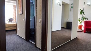 A hotel room with a bed, a desk, and a chair. There is a door leading to another room and a hallway with a plant and a red chair. - Razvan (Bucharest)