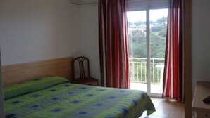 Une chambre avec un lit, une chaise, un balcon avec des rideaux rouges et une vue sur la verdure environnante.